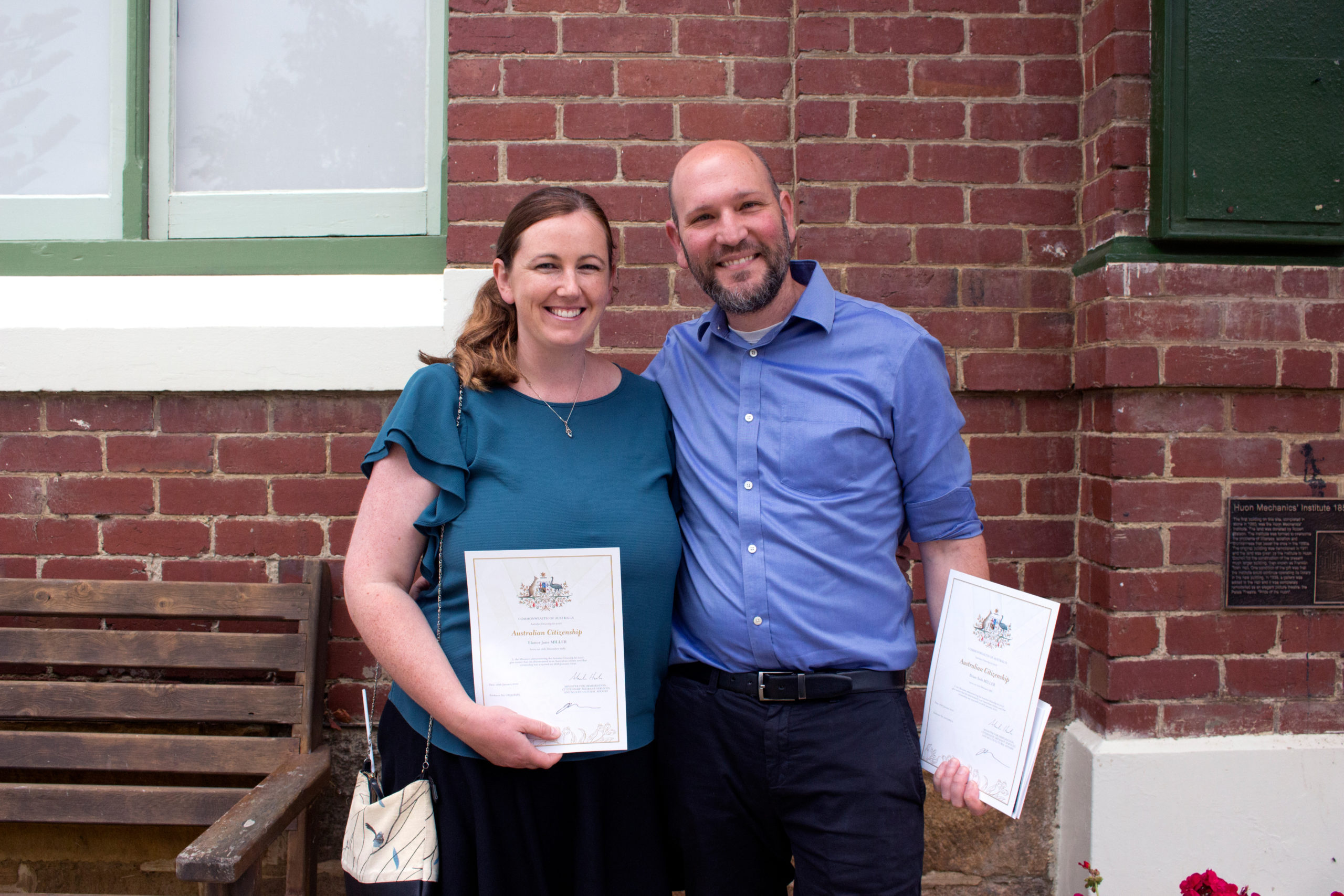 A woman and a man stand smiling in front of a brick wall, each holding a certificate. The woman is in a teal dress, and the man is in a blue shirt. They stand by a wooden bench and a plaque.