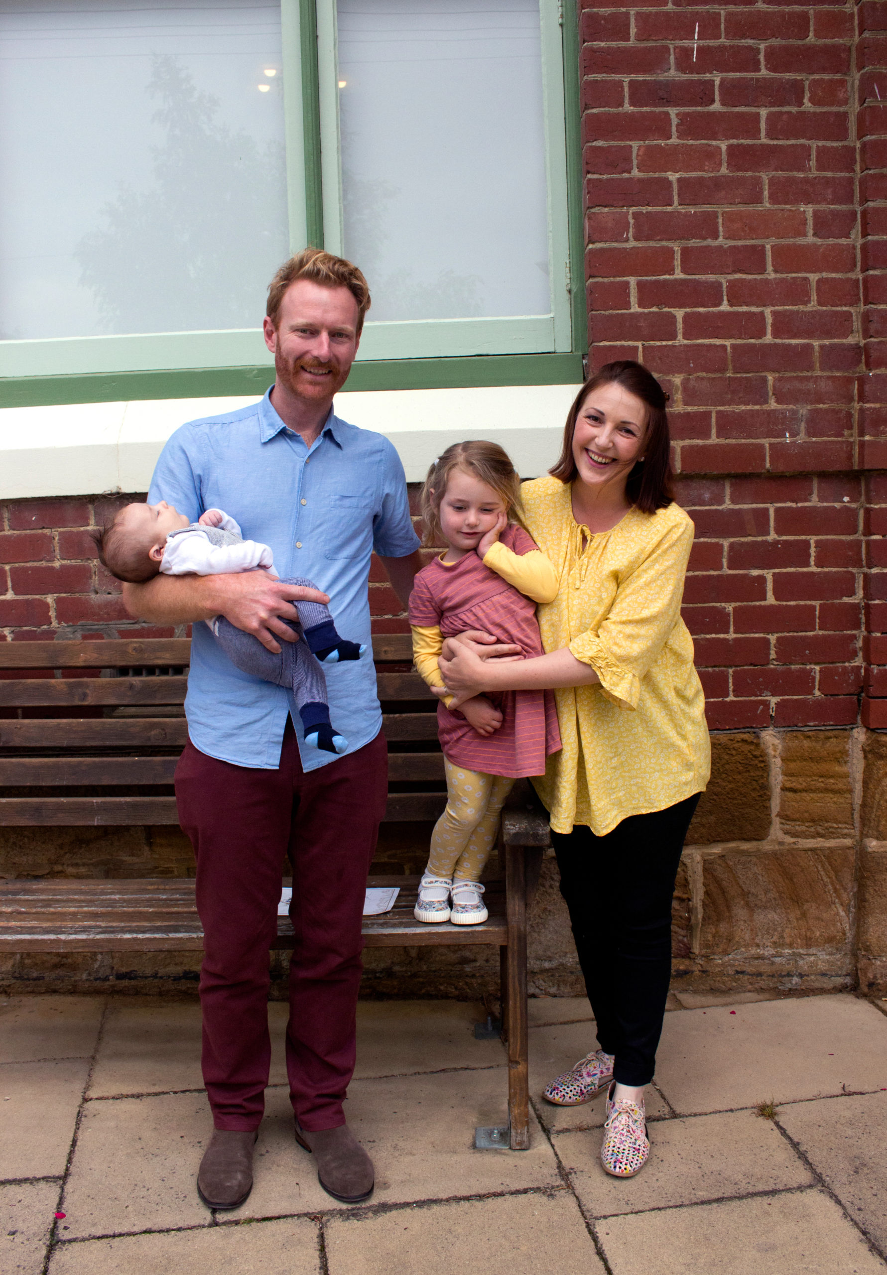 A smiling family is standing outside a brick building. The father holds a sleeping baby, while the mother holds a young girl in her arms. The father is wearing a blue shirt and the mother a yellow blouse. They stand beside a wooden bench.