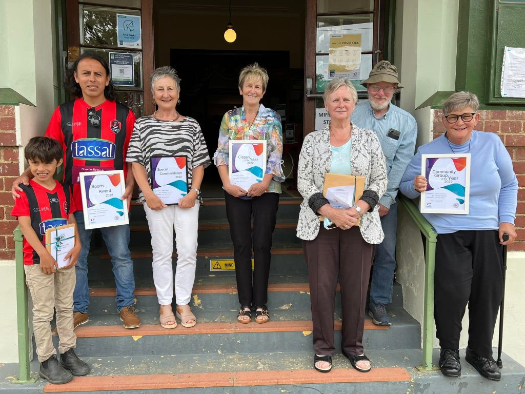 A group of seven people standing on steps outside a building, holding certificates and smiling. The group includes a child and adults, dressed casually. The entrance has posters and a lit bulb. They stand on tiled steps with a green railing.
