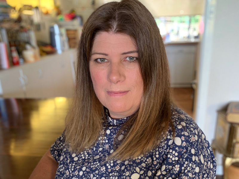 Woman with straight brown hair wearing a patterned blue and white top sits indoors, looking toward the camera with a neutral expression. A softly lit room with warm colors is blurred in the background.