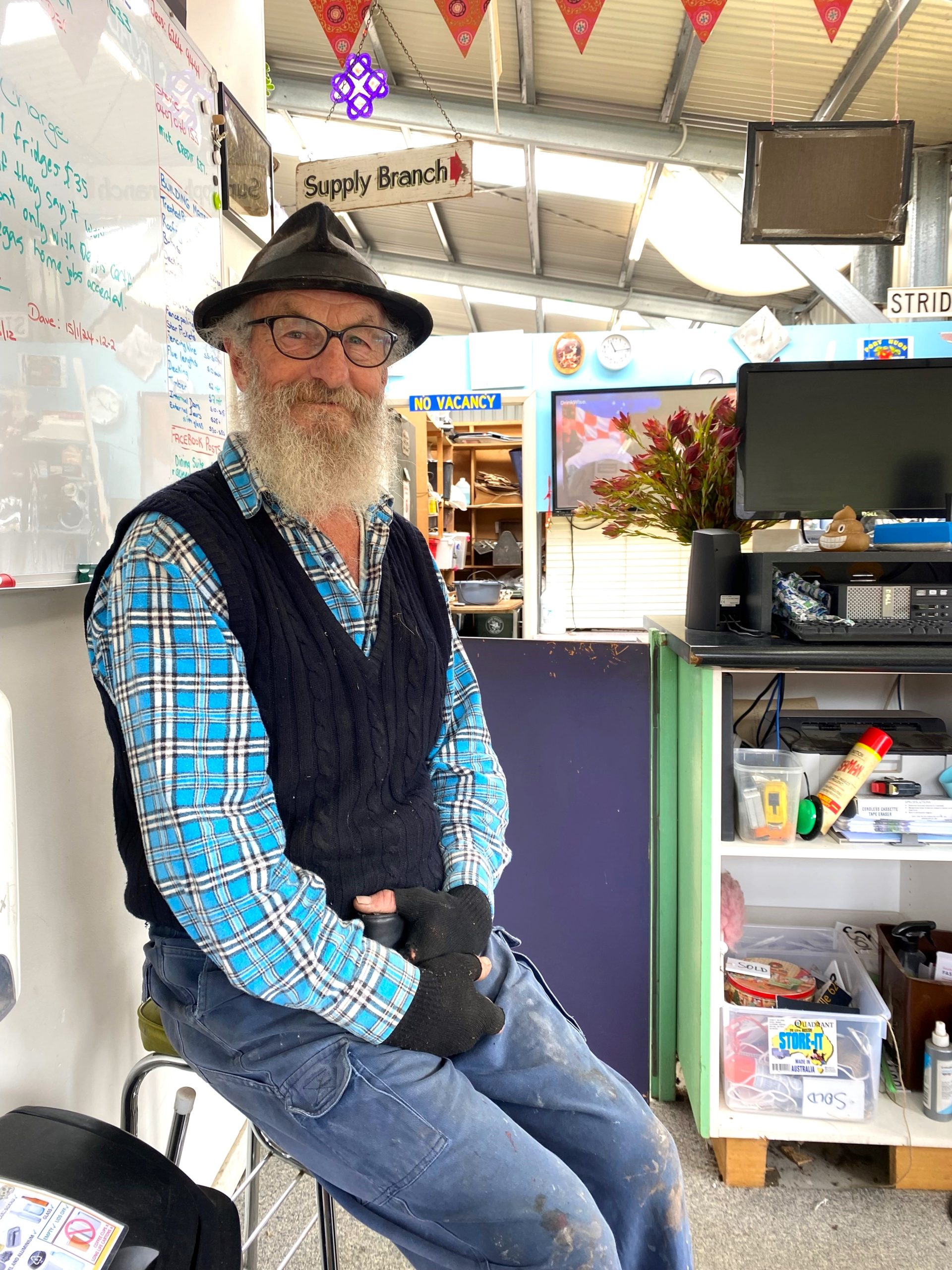 An older man with a white beard wearing a hat, blue plaid shirt, black vest, and fingerless gloves sits on a stool in a shop. Behind him are shelves with various items, a TV, and a whiteboard. A Supply Branch sign hangs above.