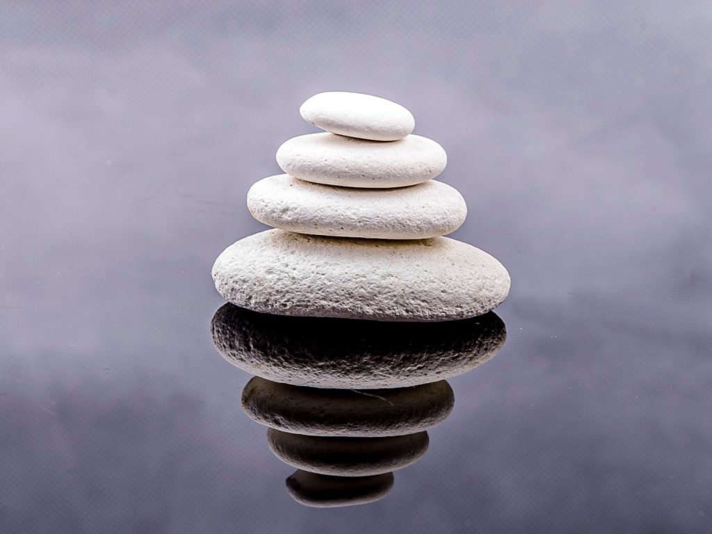 A stack of five smooth, white stones of varying sizes is balanced in a tower formation against a gray, reflective surface, creating a calming and balanced composition.