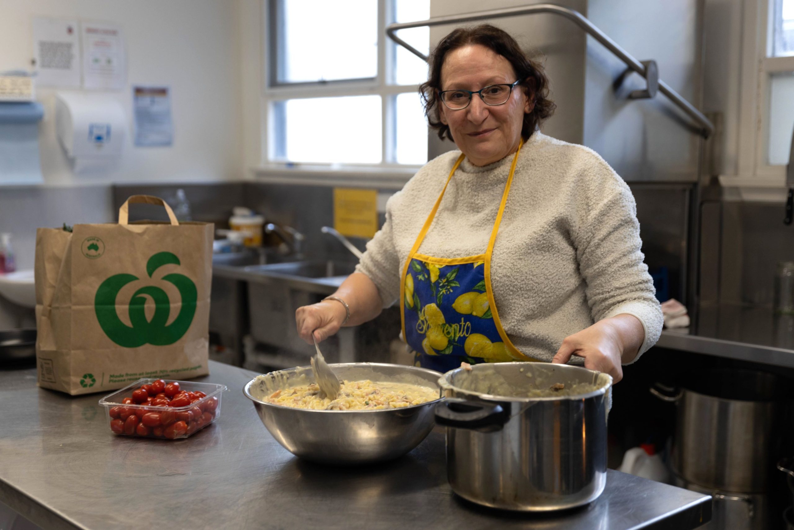 A person in a kitchen stands behind a metal countertop preparing a dish. They are wearing glasses, a gray sweater, and a colorful apron. In front of them are bowls filled with food and a bag of cherry tomatoes.