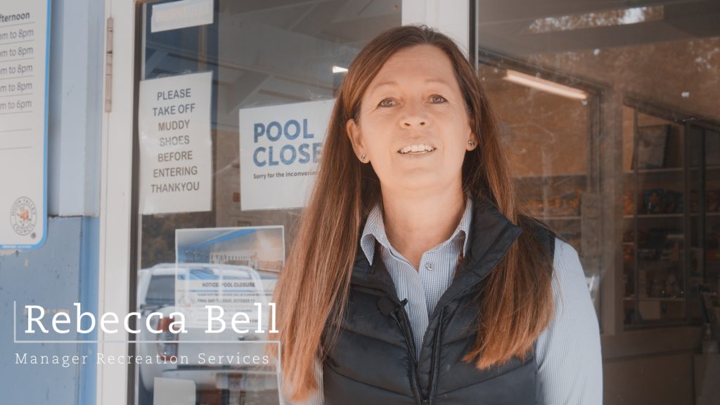 A woman stands in front of a door with a Pool Closed sign. She is wearing a blue shirt and dark vest. The overlay text reads: Rebecca Bell, Manager Recreation Services.