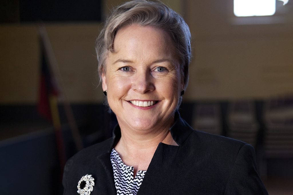 A smiling person with short gray hair, wearing a black jacket with a decorative brooch and a patterned top, stands in a dimly lit room with stacks of chairs and a flag visible in the background.