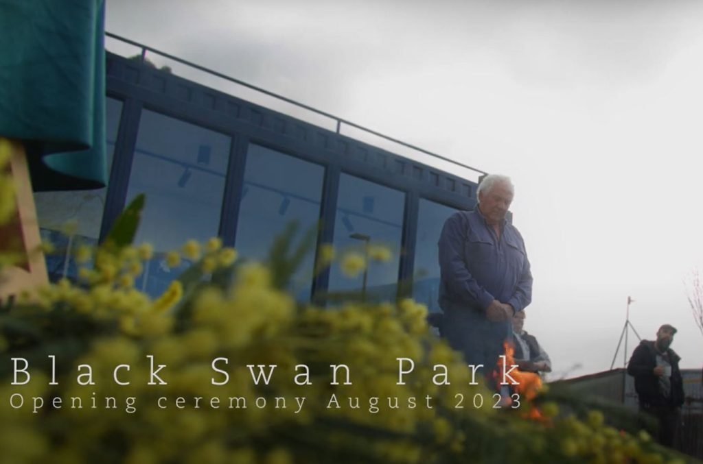 A person stands beside a fire at Black Swan Parks opening ceremony in August 2023. The background features a modern building with large windows. Yellow flowers are visible in the foreground.