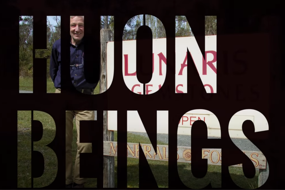 The image shows a person standing next to a sign. The words HUMAN BEINGS are boldly superimposed over the image. The person is smiling and the background includes greenery and part of the sign.