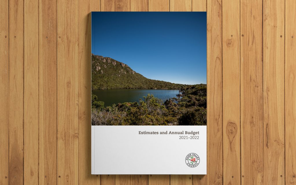 Cover of a 2021-2022 budget report on a wooden surface. The cover features a scenic landscape with a lake, green hills, and a clear blue sky, with text reading Estimates and Annual Budget 2021-2022 and a seal in the lower right corner.