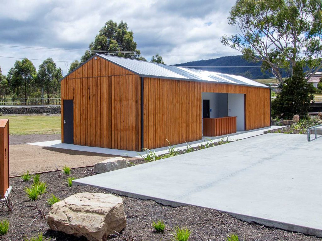 A small, modern wooden building with a metal roof stands in a landscaped area. The structure features a minimalist design with large doors and windows, surrounded by greenery and a concrete path. The sky is partly cloudy.