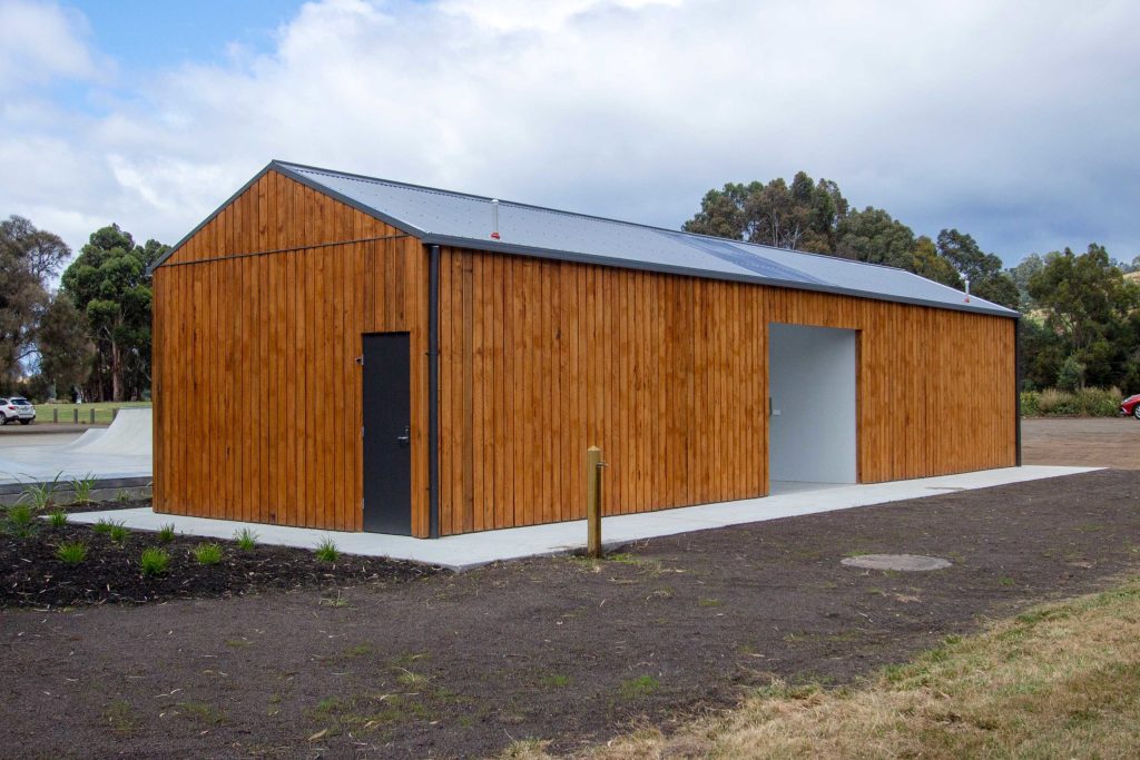 A long wooden building with a sloped roof and a small door on the left side. The structure is surrounded by grass and has a concrete path leading to it. Trees are visible in the background under a partly cloudy sky.