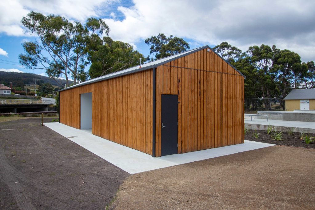 A wooden building with a sloped roof sits on a concrete foundation. It has a black door and a small window. There are trees and a cloudy sky in the background, with a gravel path leading to the entrance.