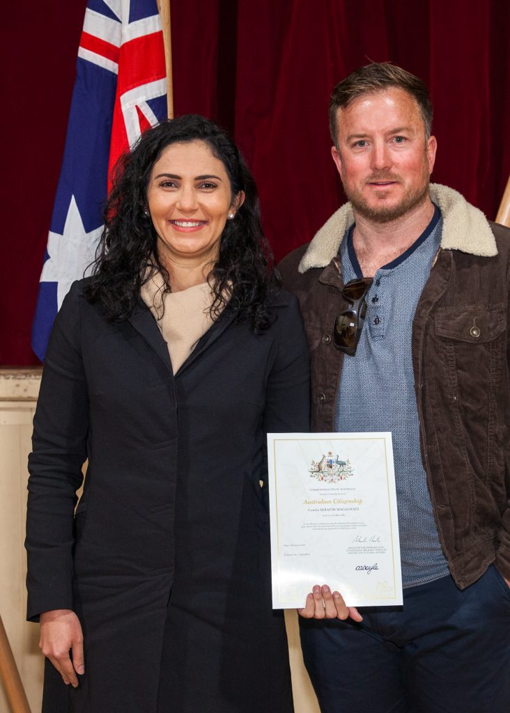 A woman and a man stand smiling. The woman holds an Australian Citizenship certificate. They are in front of an Australian flag and a red curtain.