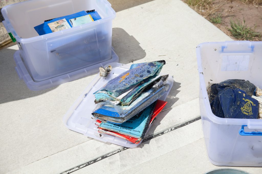 Two open plastic bins on a concrete surface, one containing stacks of weathered, water-damaged papers and booklets. Another bin is partially visible, holding various small rocks and debris.