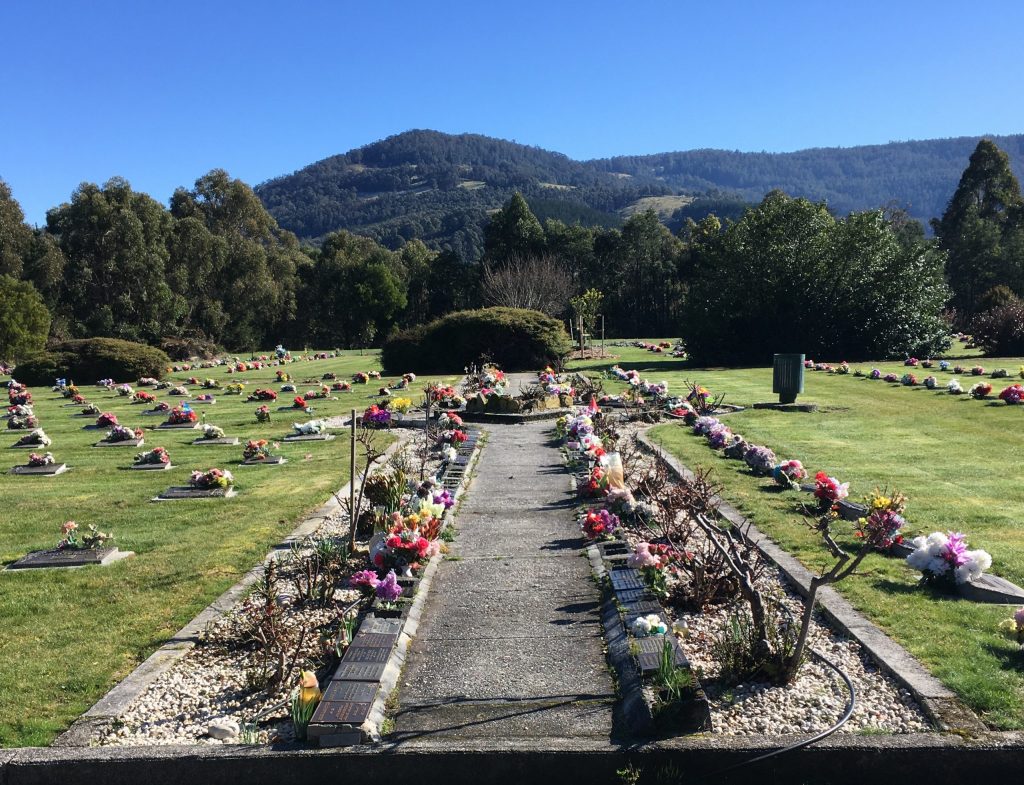 A scenic cemetery with tombstones and colorful flowers on green grass. A pathway lined with more flowers and small shrubs runs through the center. Trees and hills are visible under a clear blue sky in the background.
