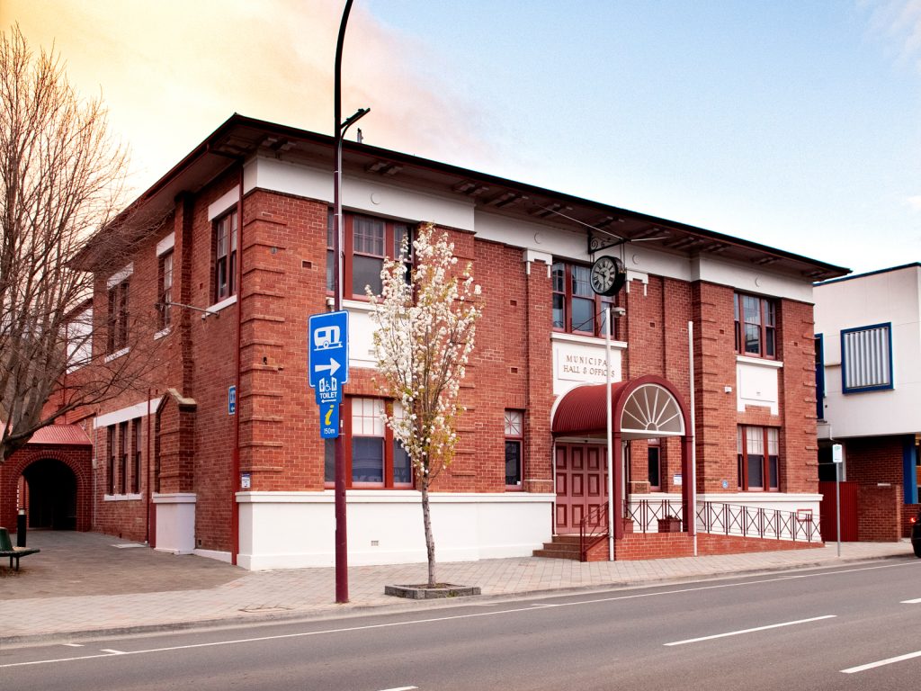 A two-story brick building with a large arched entrance awning. A clock is mounted on the front wall. The street has a blue directional sign and a few trees with sparse leaves. The sky is partially cloudy.