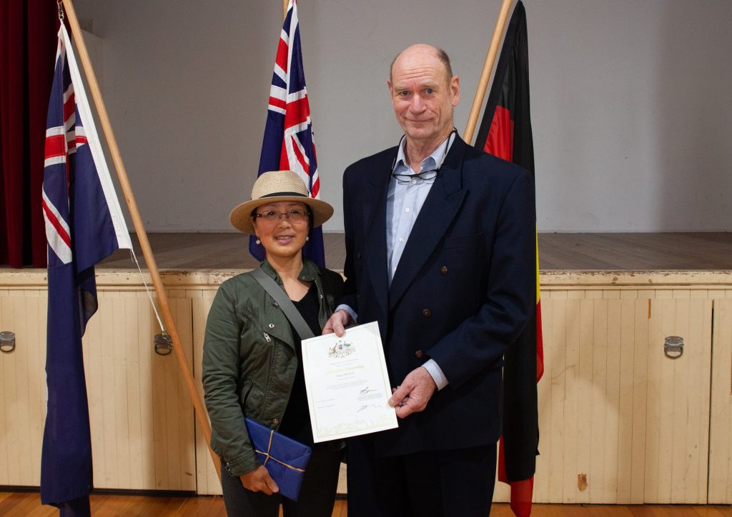 Two people stand indoors holding a certificate. One wears a hat and casual clothing, while the other is in formal attire. Three flags are behind them.