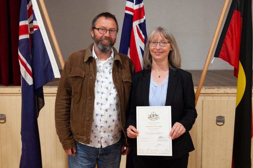 A man and a woman stand smiling. The woman holds a citizenship certificate. Behind them are three flags on stands, including Australian and Aboriginal flags. They are indoors on a stage with a wooden floor.