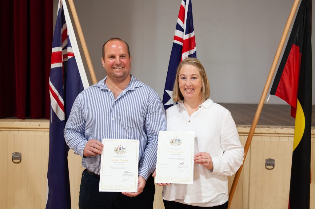 A man and a woman are smiling and holding certificates while standing in front of three flags. They are on a stage with a wooden floor and wall. The man is wearing a striped shirt and the woman is wearing a white blouse.