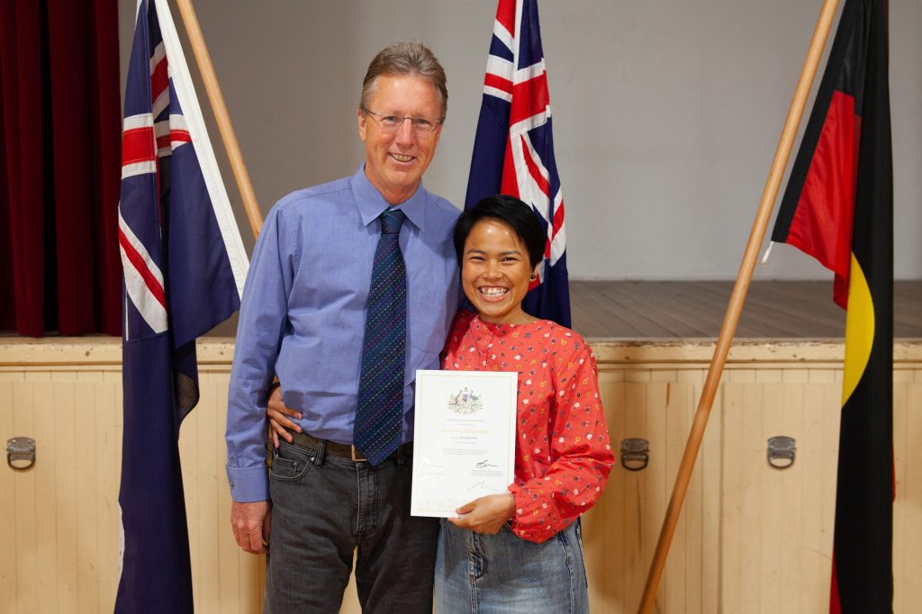 A smiling man and woman stand together in front of three flags, holding a certificate. The man wears a blue shirt and tie, while the woman wears a red, long-sleeved top with patterns. They are in a room with wooden flooring and red curtains.