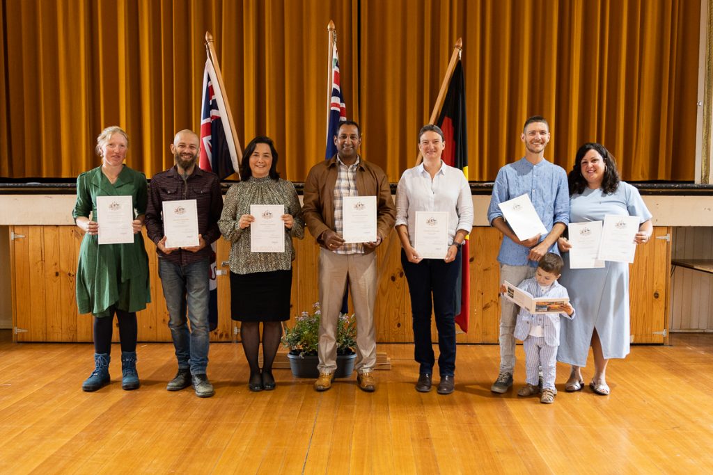 A diverse group of seven people stands on a stage holding certificates. Behind them are three flags. One person is holding a child. The setting suggests a formal or celebratory event.