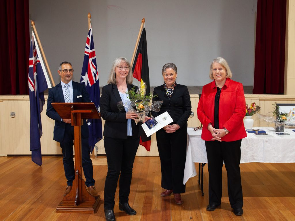 Four people stand in a hall with wooden flooring, three women and one man. One woman holds flowers and an award. Flags, including the Australian flag, and a table with more awards are visible in the background.