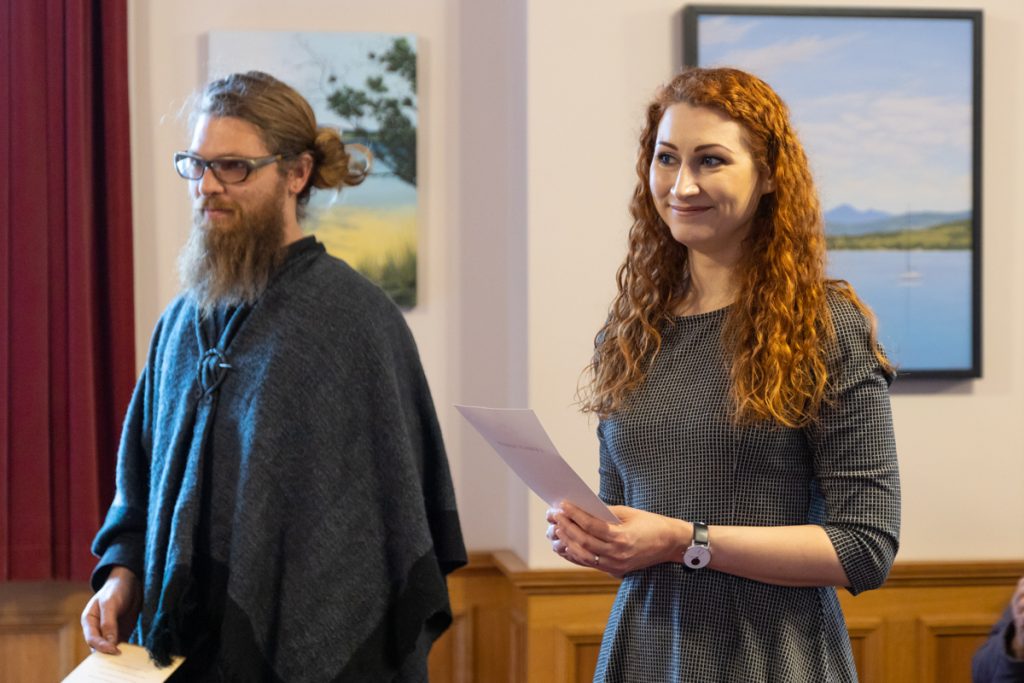 Two people stand indoors with paintings on the wall behind them. The person on the left has a beard and glasses, wearing a dark poncho. The person on the right has long red hair, wearing a plaid dress and holding papers, both appear engaged in an event.