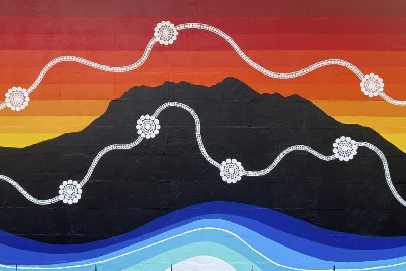A mural with a silhouette of a mountain against a sunset gradient sky of red, orange, and yellow. Wavy white floral patterns cross the sky, and blue abstract waves flow at the bottom, representing water.