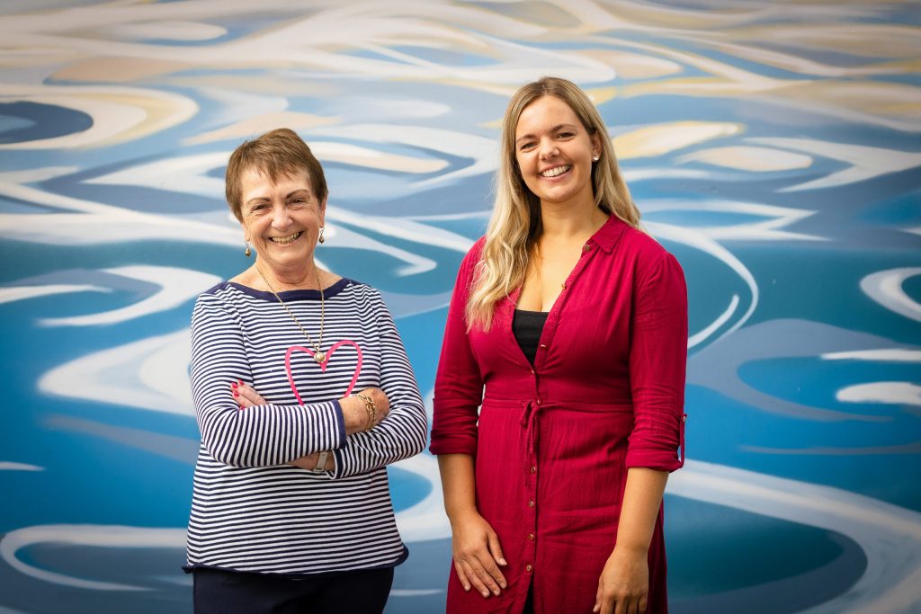 Two women smiling in front of a colorful, abstract mural with blue and white swirls. The older woman on the left wears a striped shirt, and the younger woman on the right wears a red dress. They both have a relaxed and happy demeanor.