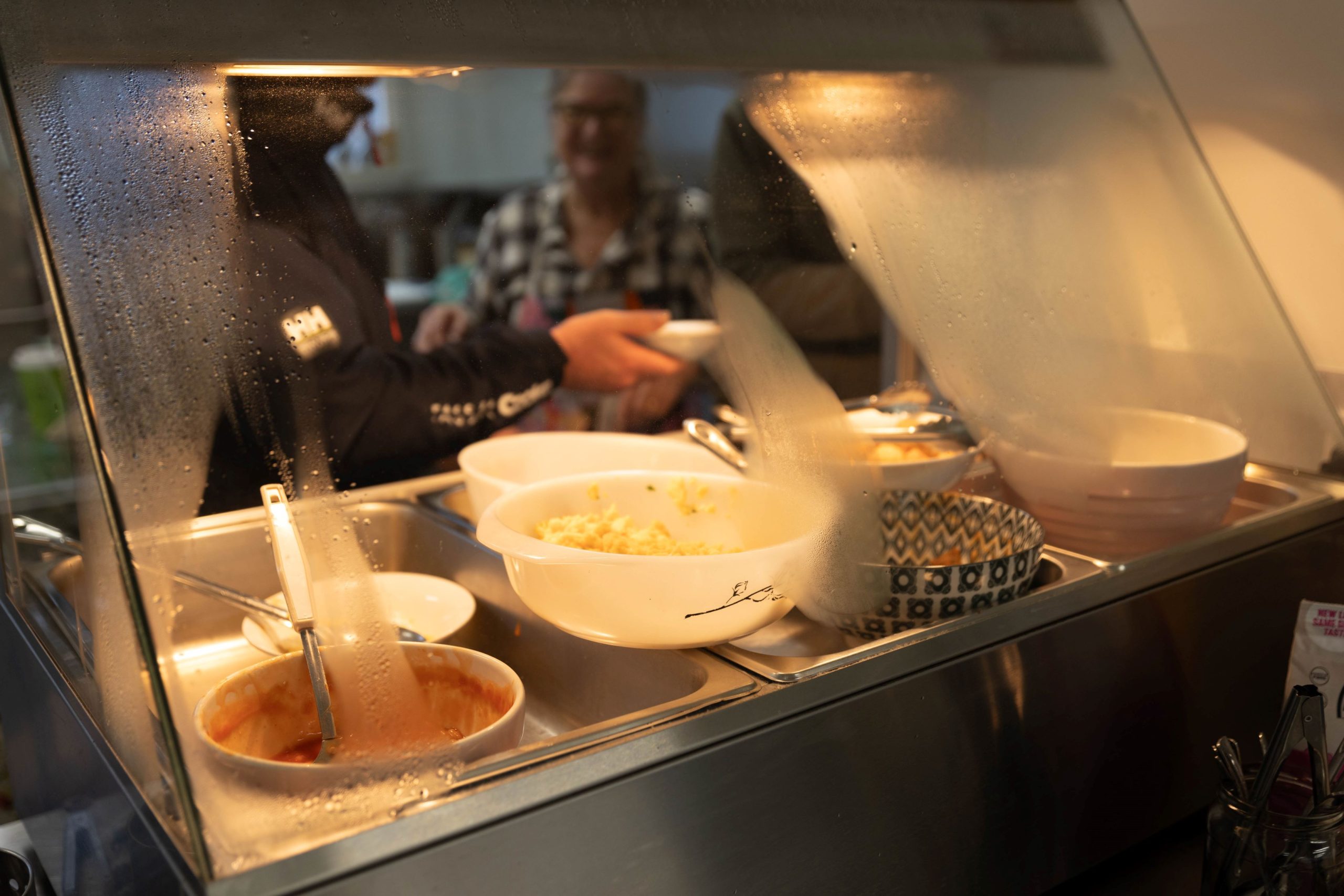 A person serves food from a buffet counter, with steam visible on the glass. Various dishes in white bowls are visible in the foreground, with a woman smiling in the background.