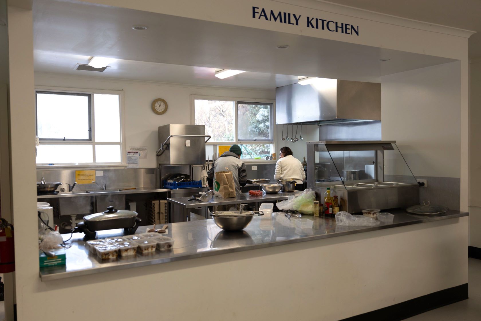 Two people work in a family kitchen with stainless steel appliances and countertops. Various kitchen tools and ingredients are on the counter. A sign above reads Family Kitchen, and there are large windows providing natural light.