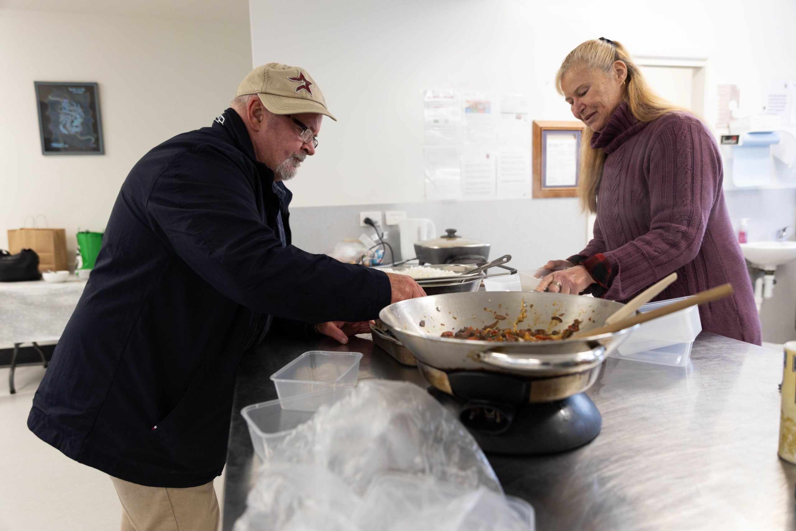 Two people in a kitchen are preparing food. A man wearing a cap and glasses is scooping food from a large pan, while a woman with long blonde hair and a burgundy sweater assists him. The kitchen counter holds various containers and utensils.