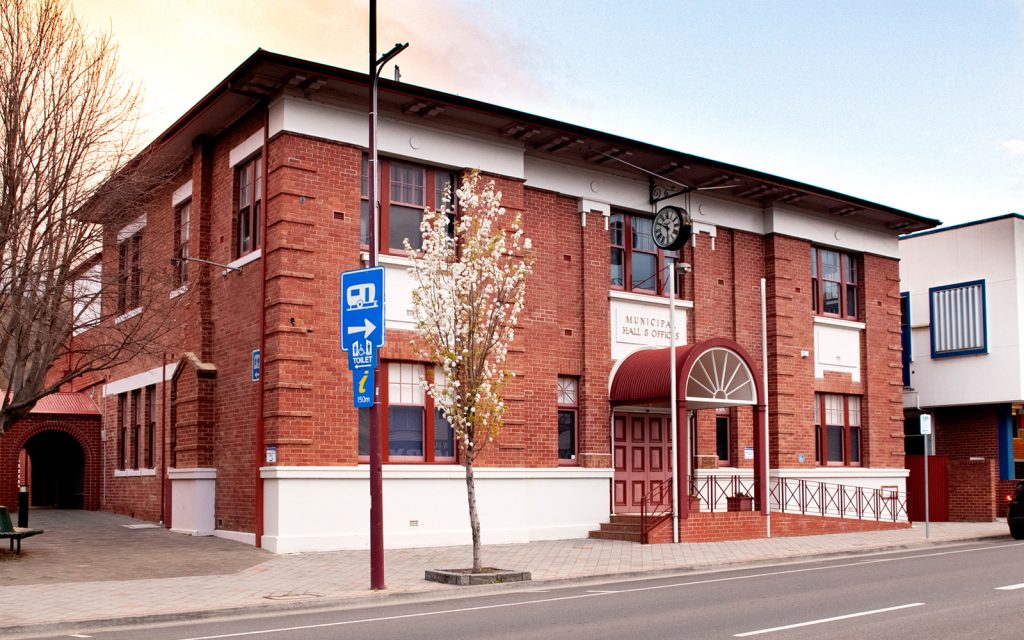 A two-story red brick building with a large arched entrance labeled Municipal Hall & Offices. It features a clock above the door, framed windows, and a small tree in front. The street is lined with signs and buildings.
