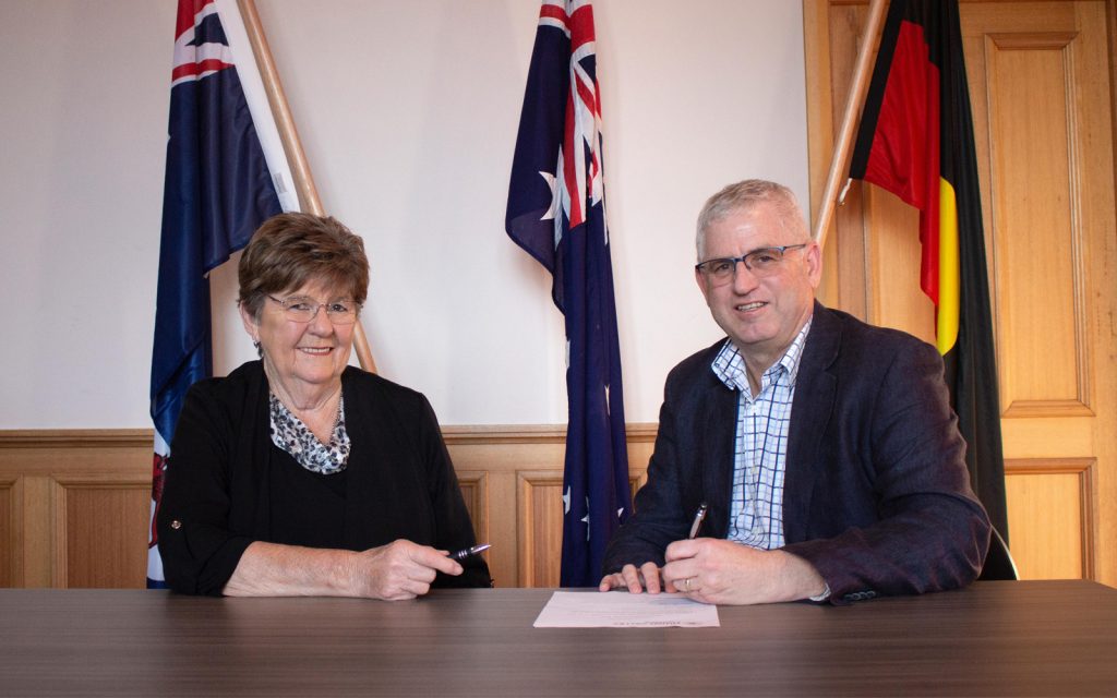 Two people sit at a table with documents, holding pens. Flags are displayed in the background, including the Australian flag. The setting appears formal, indicating an official meeting or signing event.
