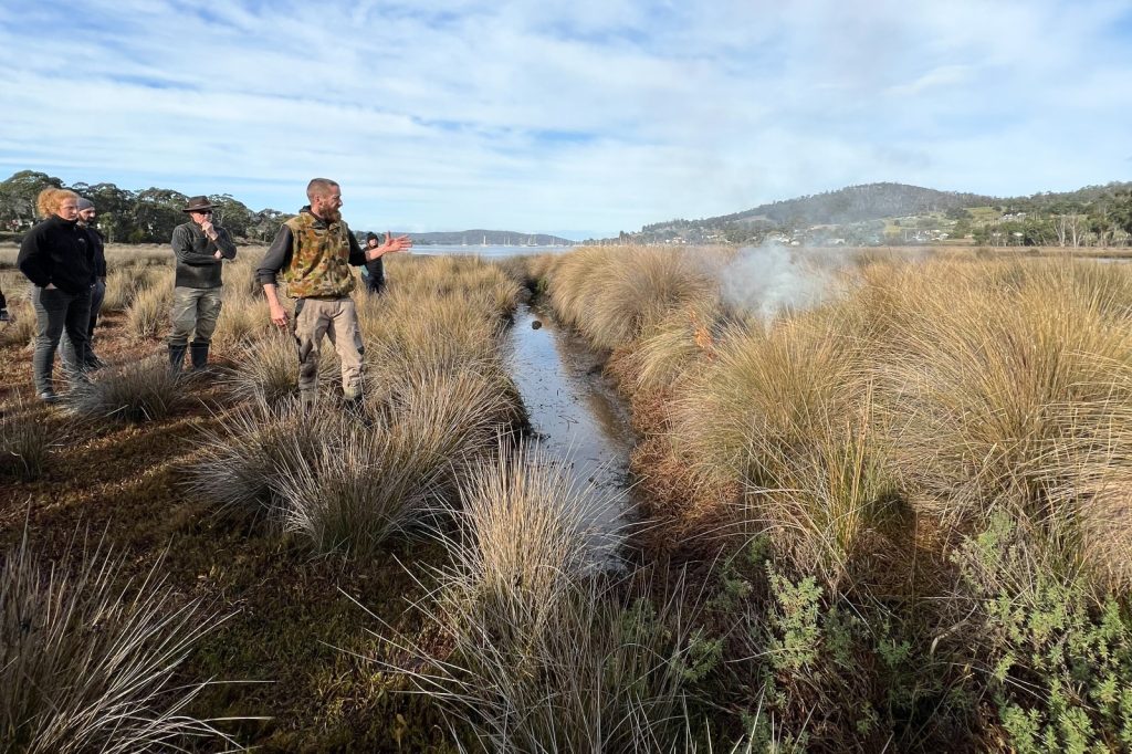 A group of people stands in a grassy wetland area. One person is gesturing towards a small controlled fire producing smoke. The landscape features dense grass, a narrow water channel, and a distant view of hills under a blue sky.