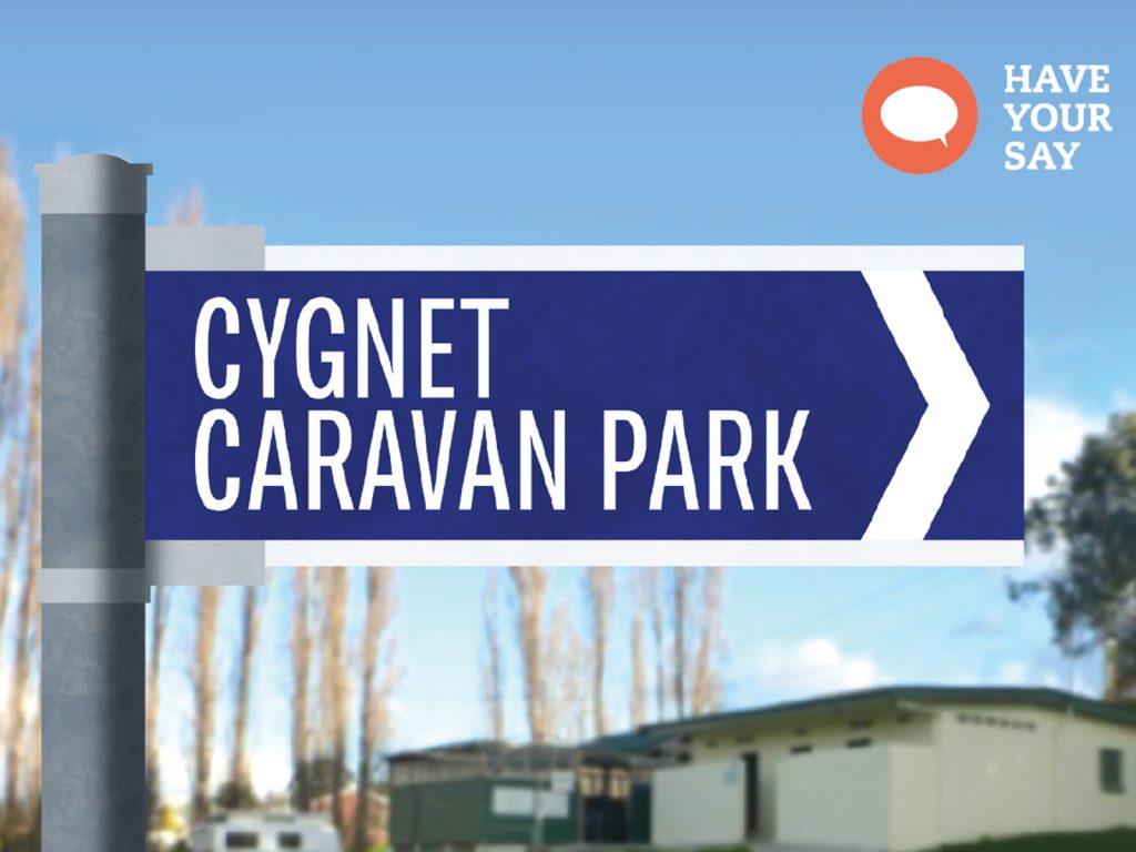 A directional sign for Cygnet Caravan Park with a blue background and white text, standing in front of blurry trees and a white building. The top right corner displays a Have Your Say logo with an orange speech bubble.
