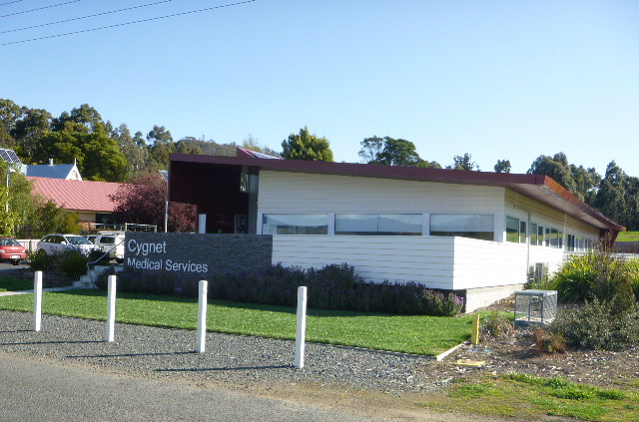 Single-story building with a modern design featuring large windows and surrounded by greenery. A sign reads Cygnet Medical Services in front. The sky is clear, and a few cars are parked nearby.