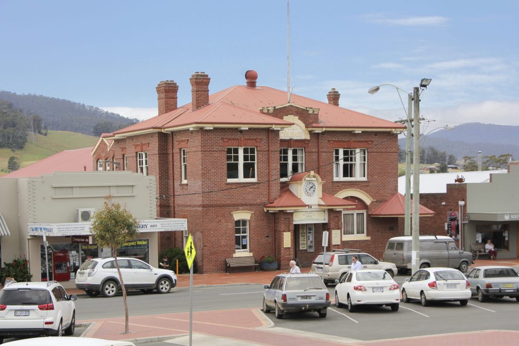 A historic two-story red brick building with a clock above the entrance. It is surrounded by parked cars and small shops. Hills and trees are in the background under a clear blue sky.