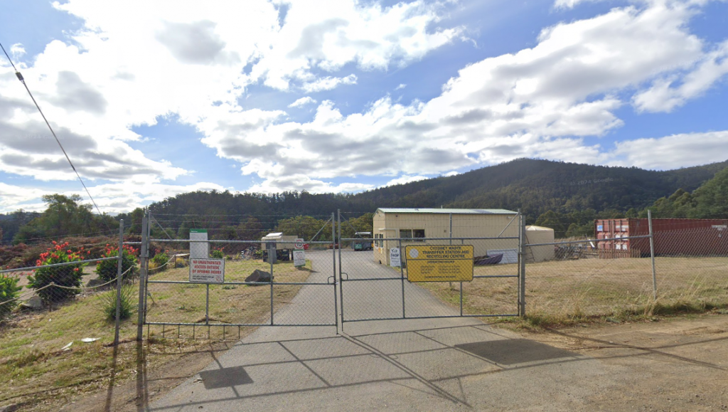 A fenced entrance leads to an industrial site with a large shed and containers. Warning signs hang on the gate. In the background, there are hills covered with trees under a partly cloudy sky.