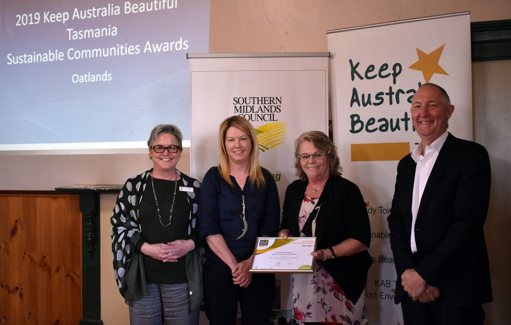 Four people stand indoors at the 2019 Keep Australia Beautiful Tasmania Sustainable Communities Awards in Oatlands. One person holds a certificate. Behind them are banners with event branding.