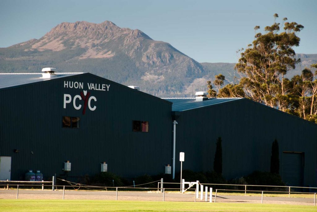 A large dark green building with HUON VALLEY PCYC written on it, set against a backdrop of a majestic mountain and clear blue sky. Tall trees are visible to the right, and a foreground of grass and fencing lines the scene.