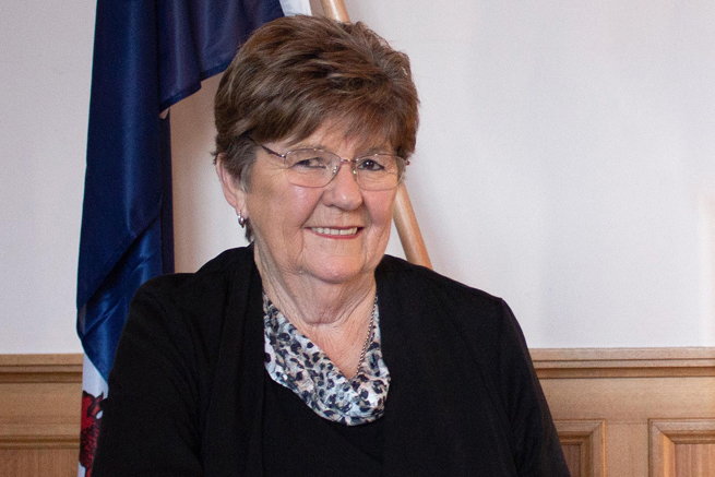 An older woman with short brown hair and glasses is smiling. She is wearing a black top with a patterned scarf. Behind her, there is a blue flag and a wood-paneled wall.