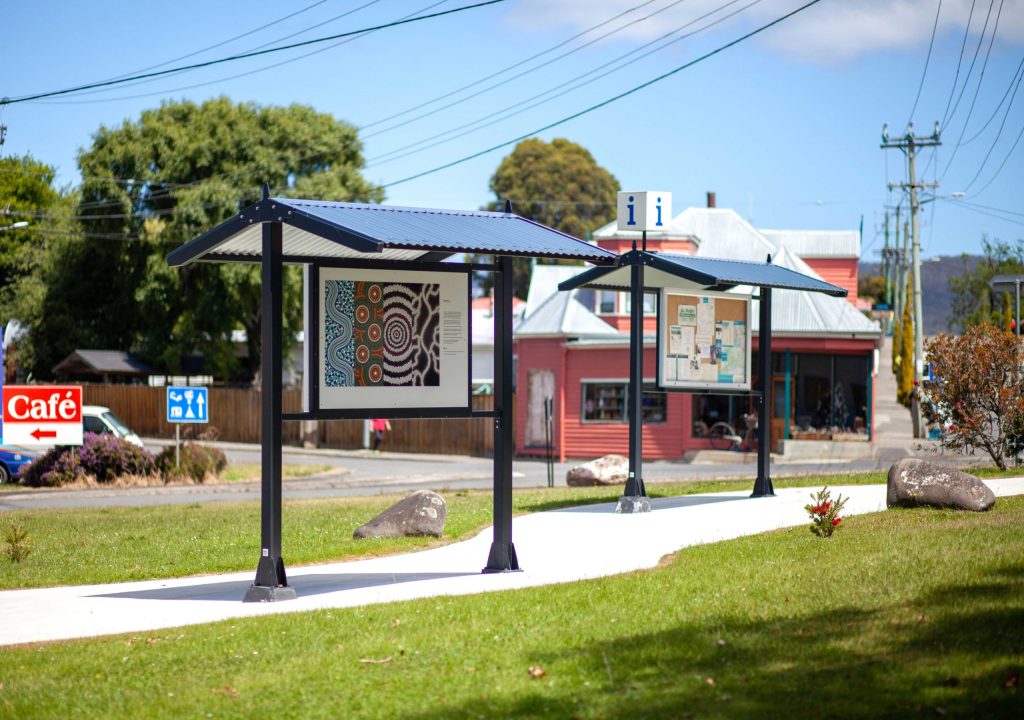 Outdoor bulletin boards with a corrugated metal roof display community notices and local art. Located on a grassy area near a café and other small buildings, with trees and power lines in the background.