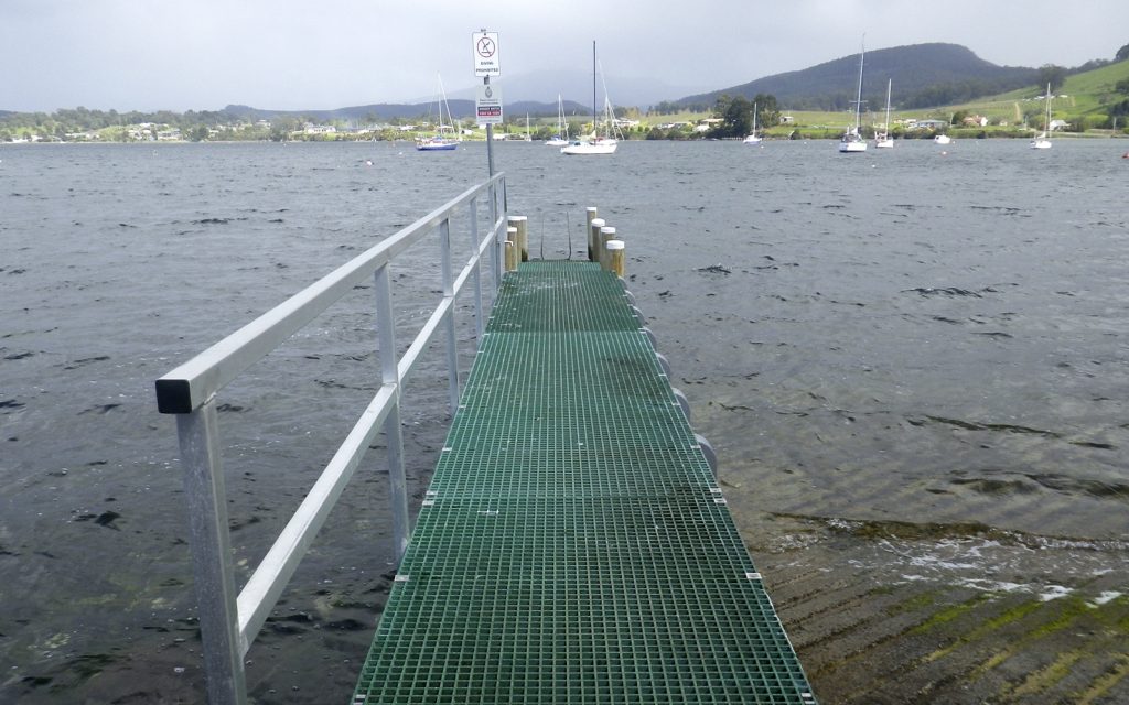 The image shows a narrow green metal boardwalk leading to a jetty over a large body of water, with several moored boats visible in the distance. Hills and a cloudy sky are in the background.