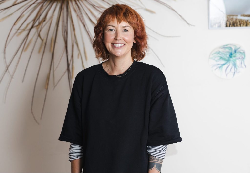A smiling person with short, wavy red hair stands in a room with white walls, wearing a black shirt over a striped top. Decorative elements, including a starburst design, are visible in the background.