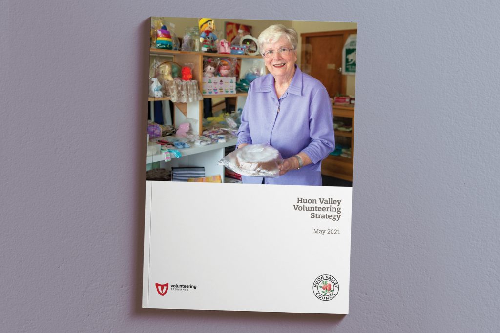 Cover of the Huon Valley Volunteering Strategy brochure, May 2021. It features a smiling elderly woman in a purple shirt holding a cake, standing in a room filled with shelves of colorful items. Logos are at the bottom.