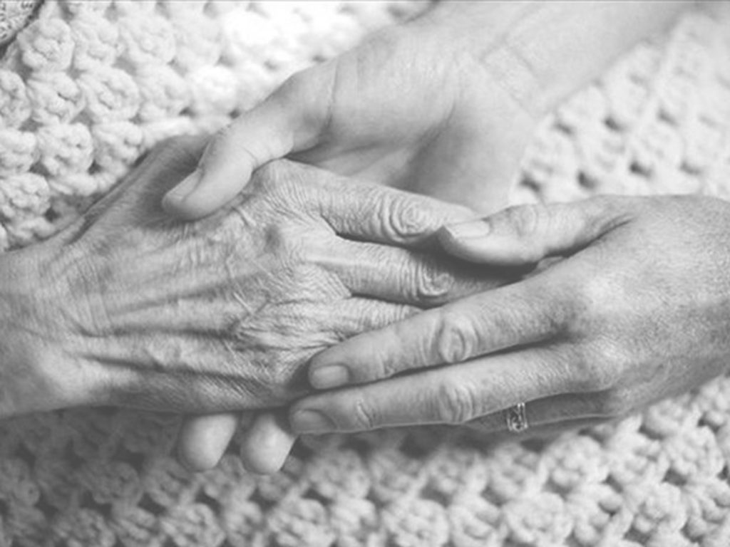 Close-up of a pair of young hands gently holding an older persons hand, both resting on a knitted blanket. The image is in black and white, highlighting the contrast between the smooth and wrinkled skin.
