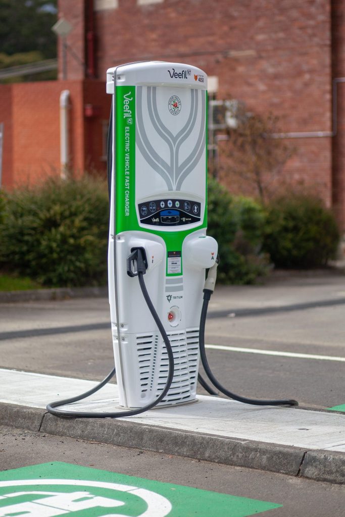 An electric vehicle charging station with two connectors is situated on a paved area. The unit features a green and white design and is installed near a red brick building and some greenery.