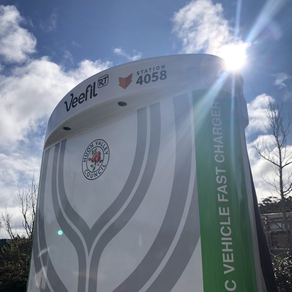 Electric vehicle fast charger with Veefil RT branding and Station 4058 text. It features a logo for Huon Valley Council. The sky is partly cloudy with the sun peeking through.