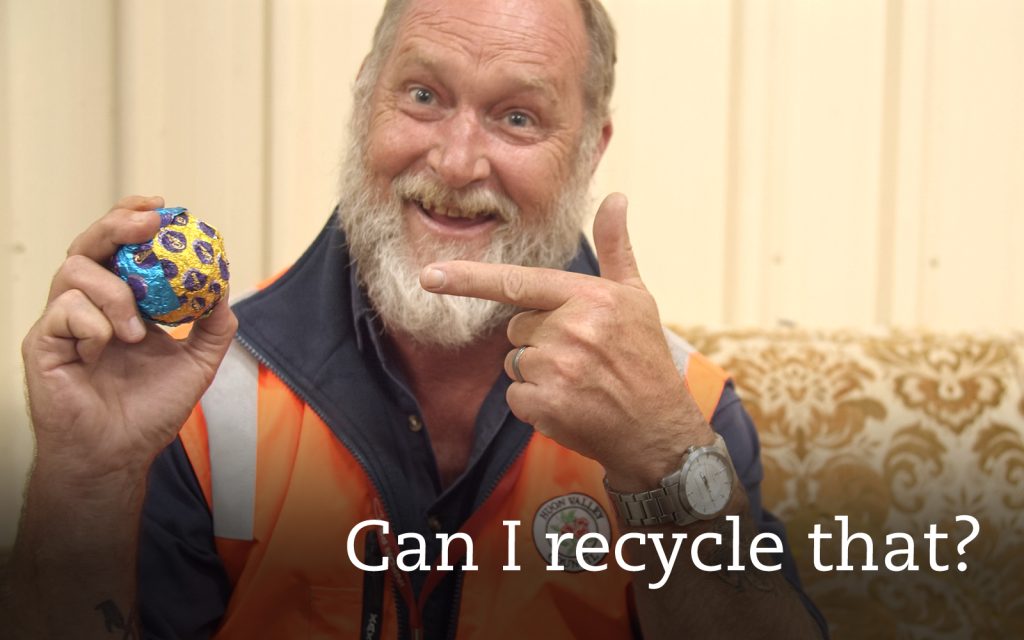 A man with a beard and wearing an orange safety vest smiles and points at a colorful, decorated ball he is holding. The text on the image says, Can I recycle that?.