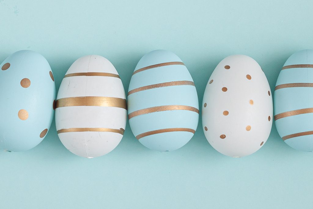 Five pastel blue and white Easter eggs arranged in a row, each with different gold patterns including dots and stripes, set against a light blue background.